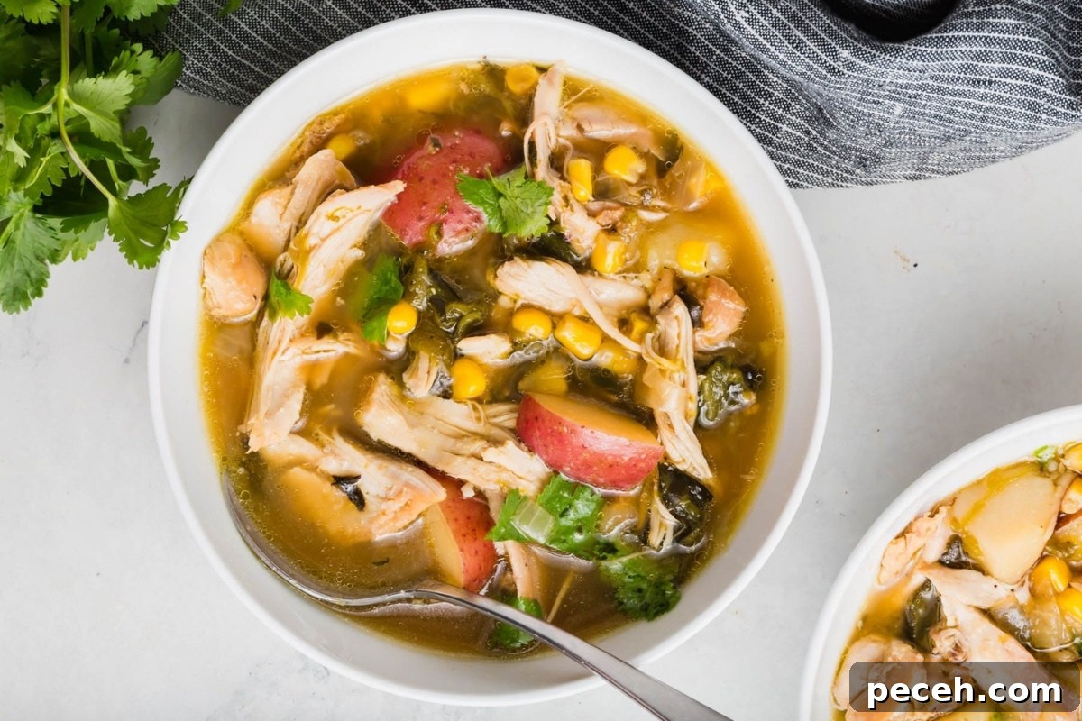 Roasted Chicken and Poblano Soup Recipe 16 A bowl of poblano chicken soup with shredded chicken, corn, red potatoes, spinach, and fresh cilantro, served with a spoon on a white marble surface next to a gray cloth napkin and fresh cilantro.