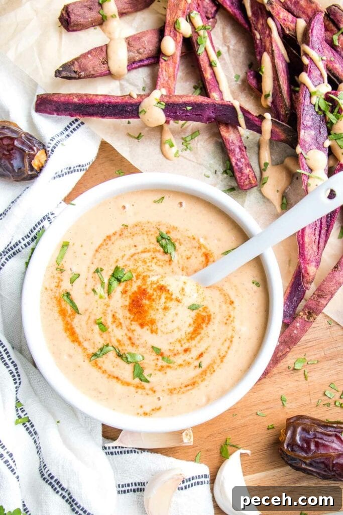 Smoky Chipotle Salad Dressing Recipe: Zesty, Creamy, & Homemade 2 small white bowl with chipotle salad dressing, swirled with extra chipotle powder and chopped cilantro, surrounded by purple fries, dates, garlic, and a white dish towel with thin blue lines. Fries are drizzled with the creamy orange dressing.