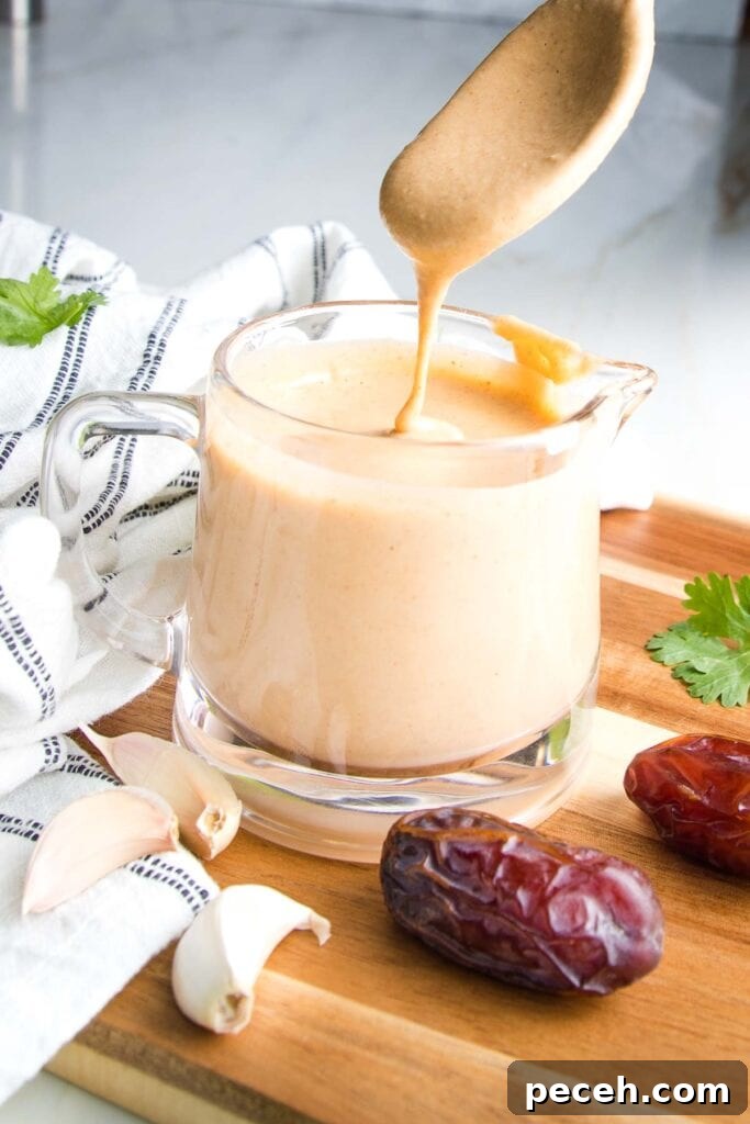 Smoky Chipotle Salad Dressing Recipe: Zesty, Creamy, & Homemade 7 Glass pitcher filled with creamy light orange dressing. There is a spoon dripping dressing back into container...a few dates and cloves of garlic on the wooden board, dish towel or countertop surrounding the salad dressing.