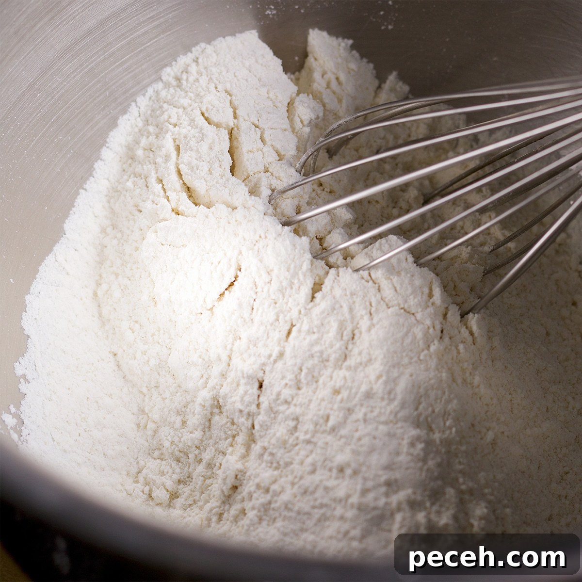 Someone using a wire whisk to mix flour and sugar in a mixing bowl.