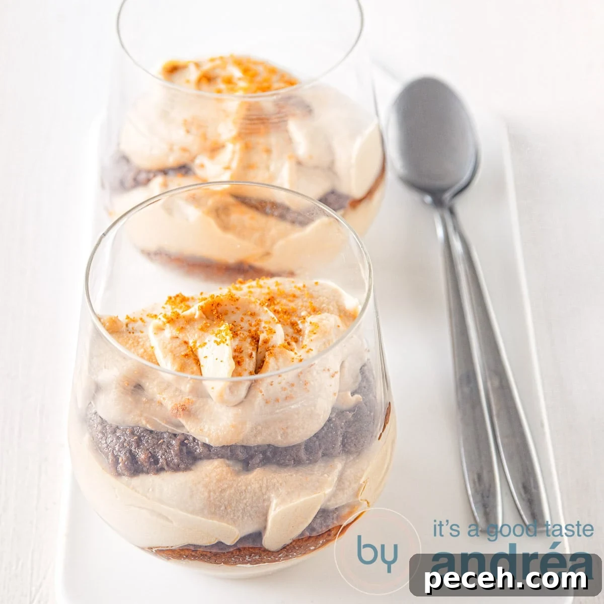 Speculoos Tiramisu Recipe with Biscoff Spread 4 A square photo with two glasses of speculoos tiramisu behind each other on a white plate. Two spoons are next to it on the right.