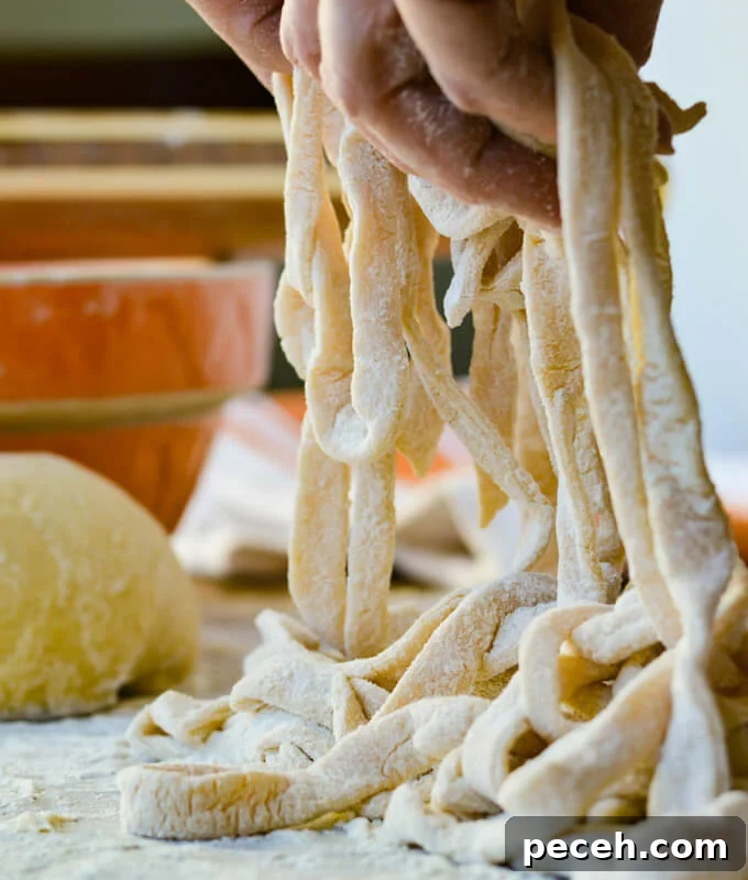 Uncooked homemade egg noodles held up with a hand. An orange dish and ball of dough are in the background.
