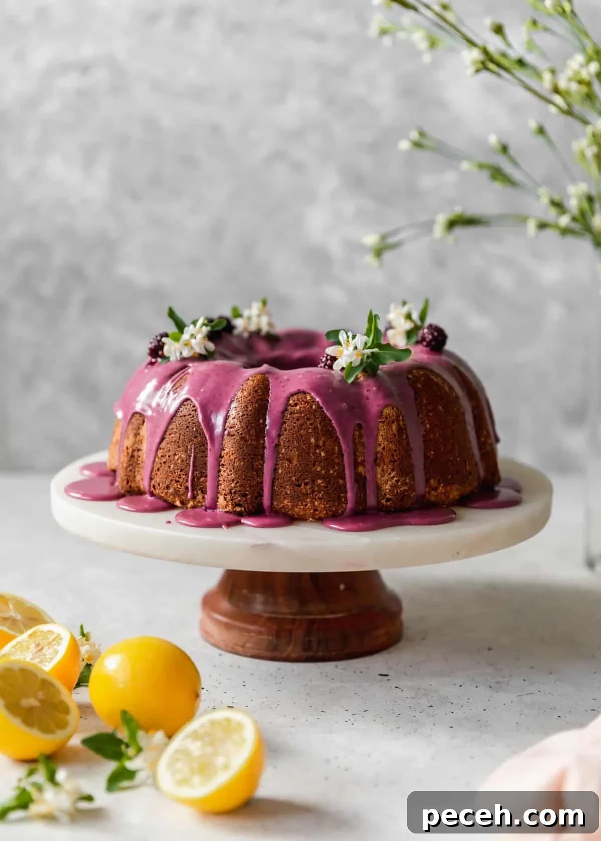 Lemon Sour Cream Pound Cake with Blackberry Glaze 2 A side image of a bundt cake topped with pink glaze and white blossoms on a marble cake stand placed on a grey background next to sliced lemons.