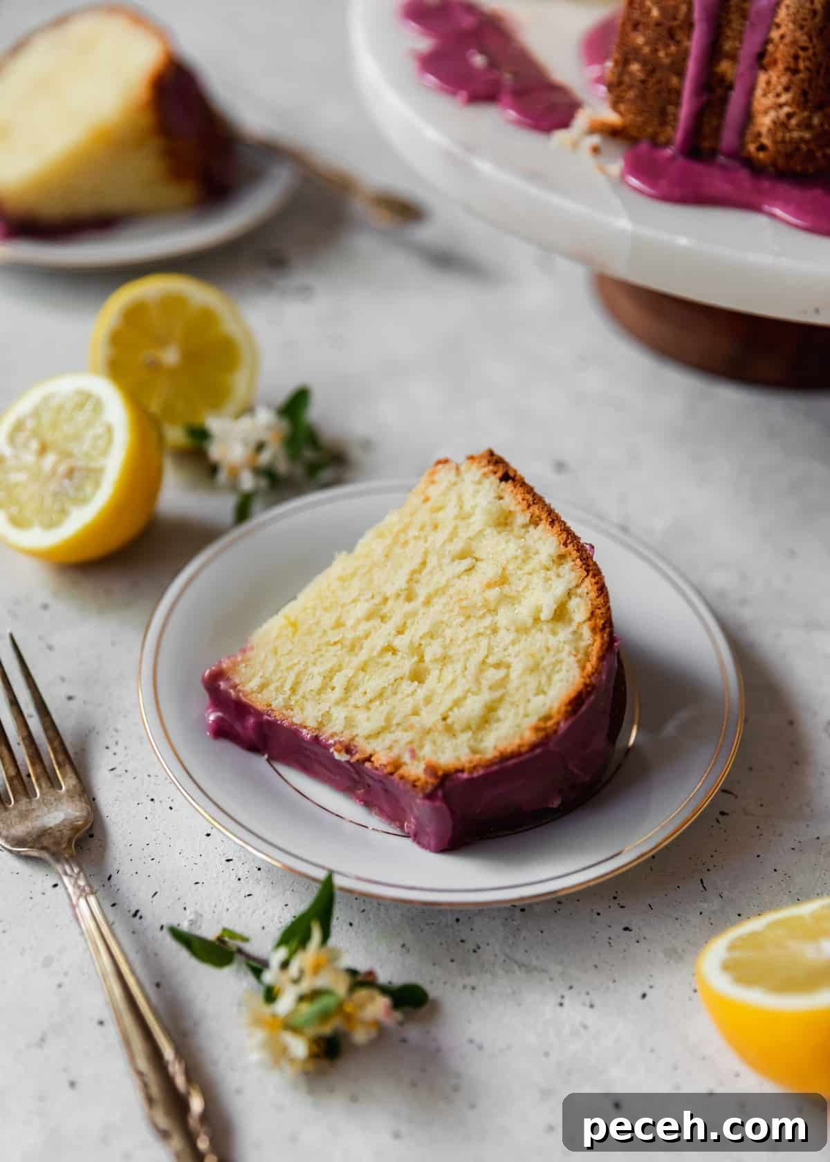 Lemon Sour Cream Pound Cake with Blackberry Glaze 3 A closeup side image of a slice of cake with pink glaze on a white plate placed on a grey table next to a fork, halved lemons, and white flower blossoms.