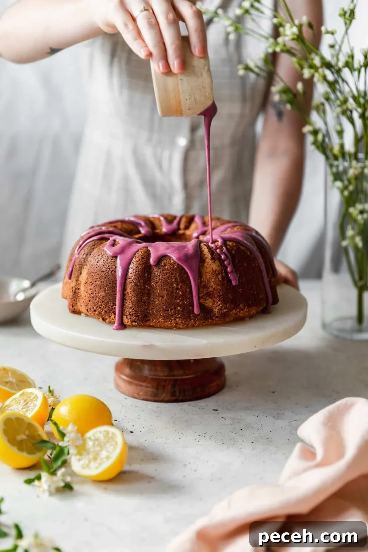 Lemon Sour Cream Pound Cake with Blackberry Glaze 7 A side image of a woman wearing a white dress pouring blackberry glaze over sour cream lemon pound cake, placed on a marble cake stand next to lemons, a pink linen, and green blossoms with a grey background.