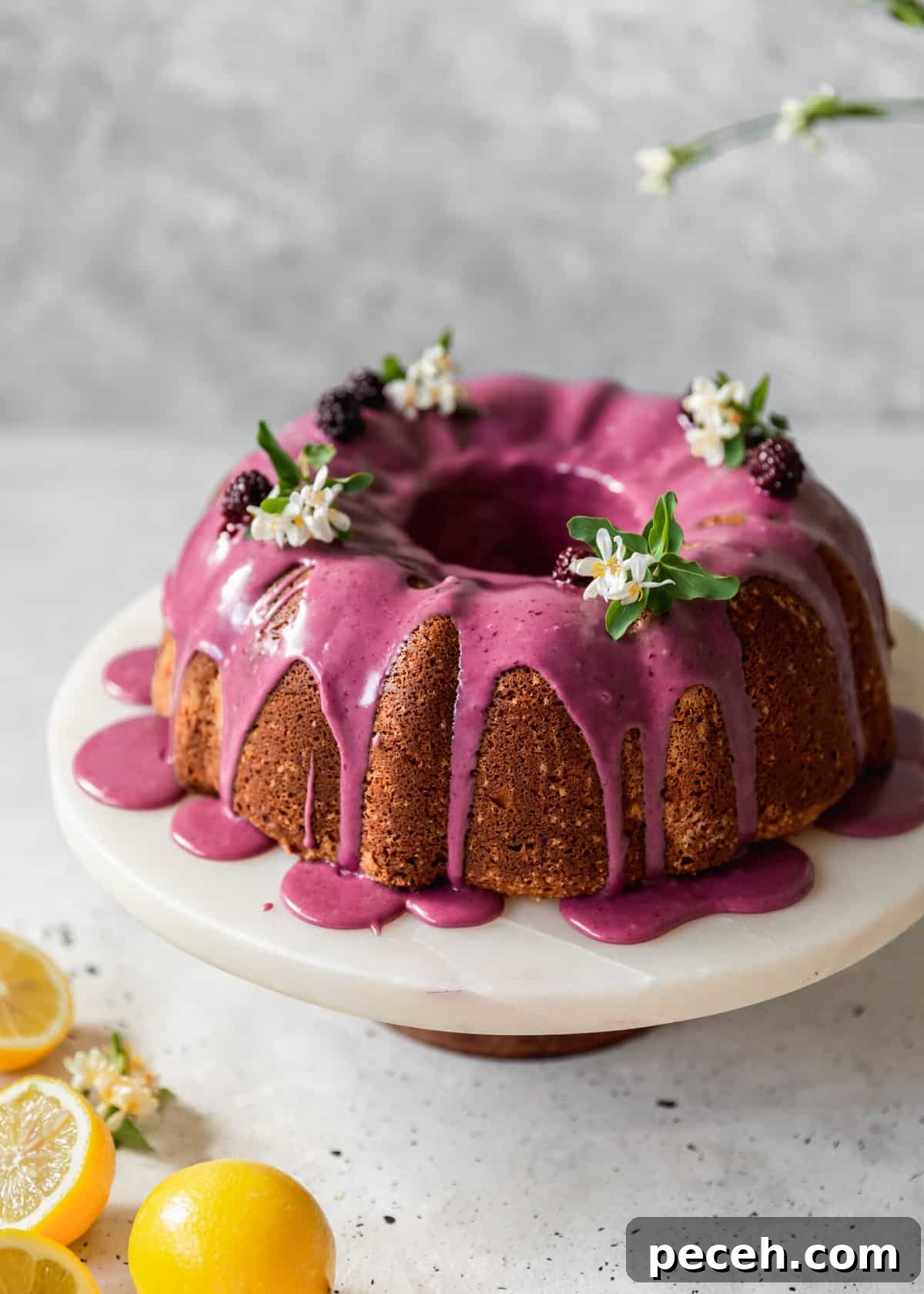 Lemon Sour Cream Pound Cake with Blackberry Glaze 8 A closeup side image of a sour cream lemon pound cake with blackberry glaze on a marble plate next to some sliced lemons on a grey table.
