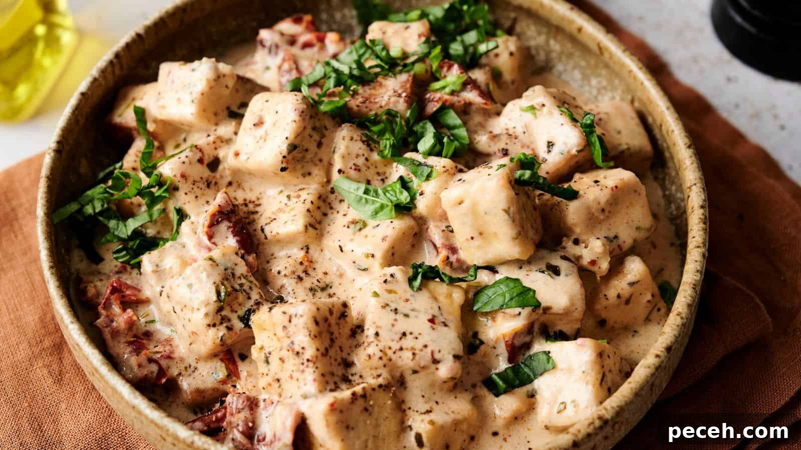 A bowl of creamy pasta salad with cubed noodles and sun-dried tomatoes, garnished with fresh basil leaves and black pepper, served in a rustic dish on a brown cloth.