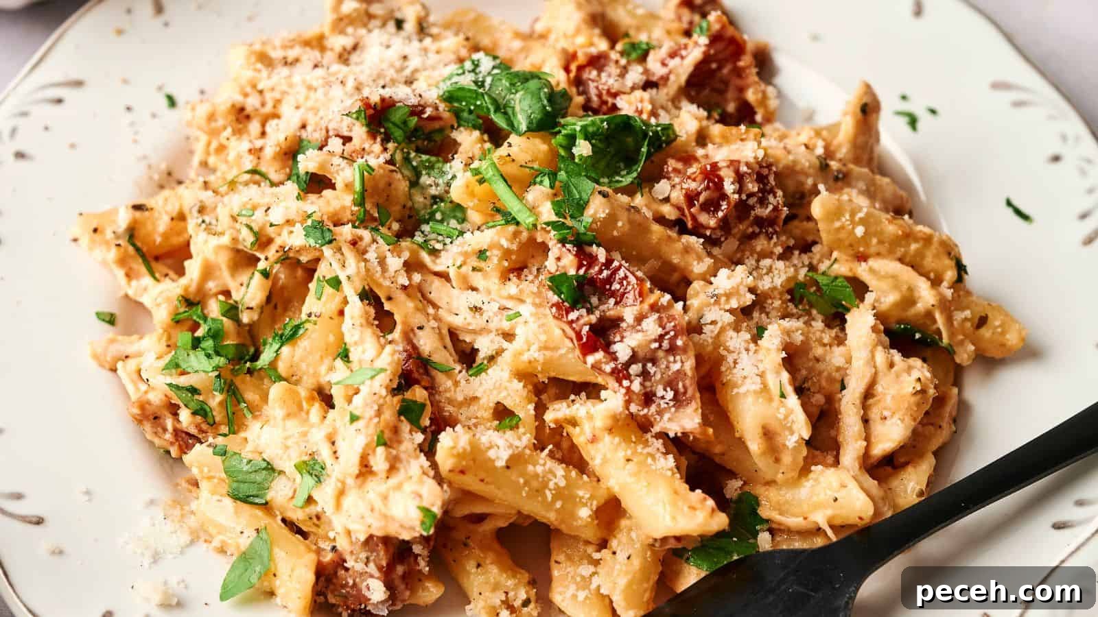 A plate of creamy penne pasta garnished with chopped parsley and grated cheese, featuring pieces of sun-dried tomatoes. A black fork is resting on the plate.