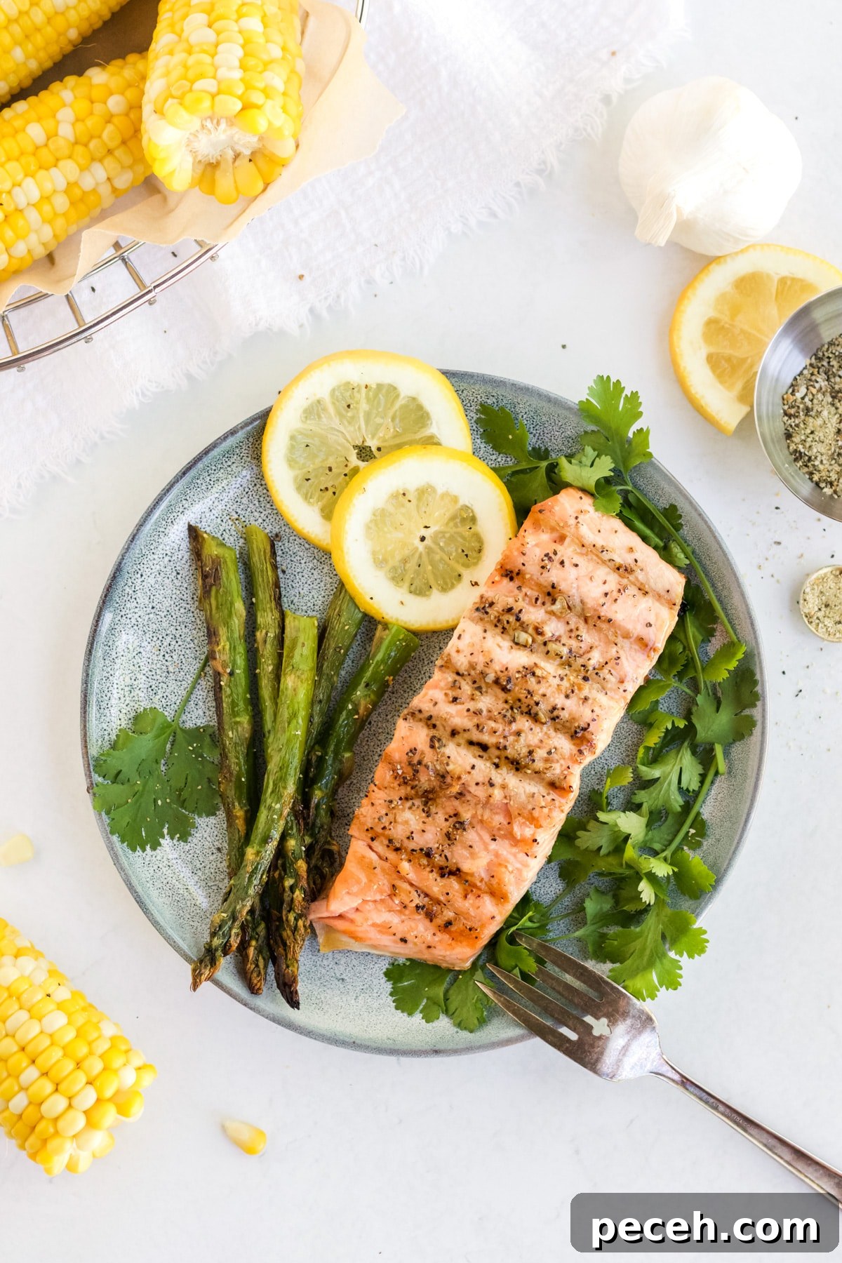 Perfectly Grilled Salmon with Crispy Skin and Lemon Herb Rub 6 Salmon filet on a plate with herbs, asparagus and lemon, corn on the cob is off in the corner.