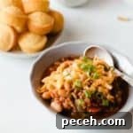 a bowl of chili with cheddar cheese and green onions on top. Mini cornbread muffins in background.
