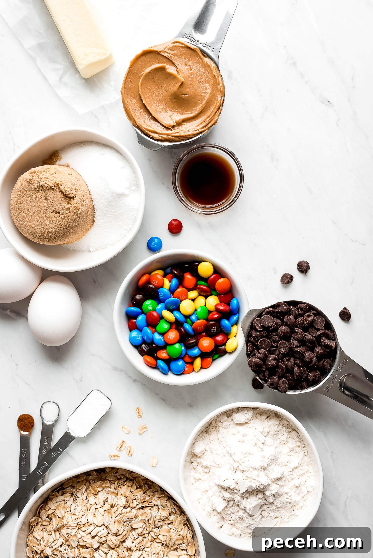 Soft Chewy Monster Cookies Recipe – Loaded Chocolate Chip Cookies 5 Measuring cups and bowls of peanut butter, sugar, eggs, M&M's, chocolate chips, oats, flour, vanilla, baking soda, cinnamon, and salt.