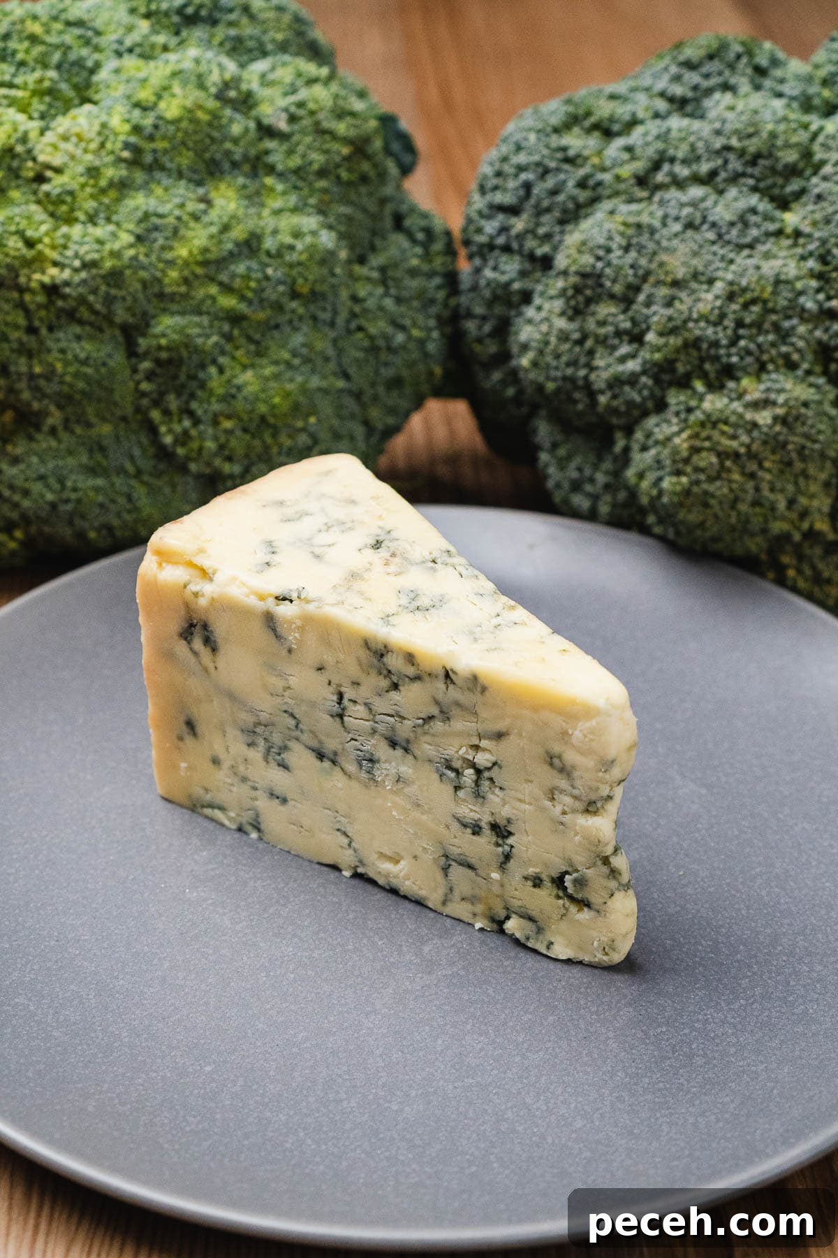 A wedge of stilton cheese on a grey plate and two heads of broccoli in the background
