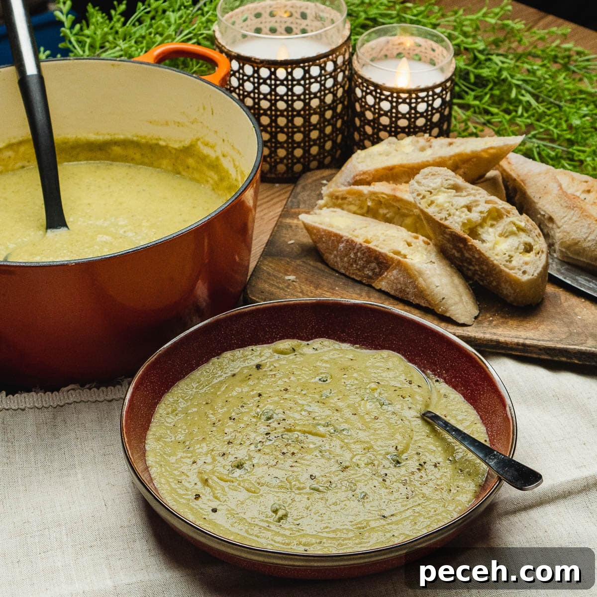 Broccoli and Stilton Soup served in a red bowl with a spoon and in the background there is crusty bread,candles and a cooking pot full with soup