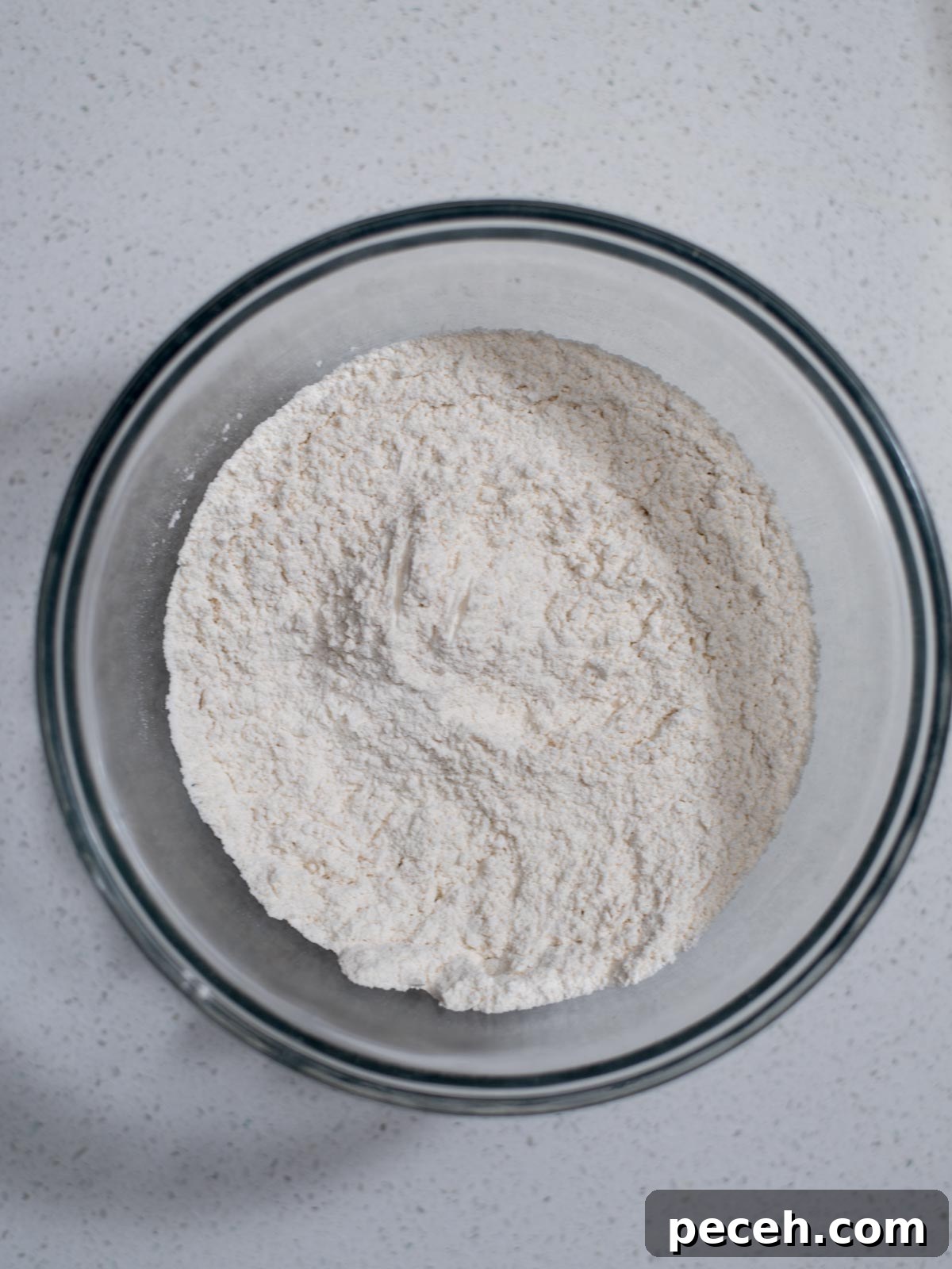 Classic Old-Fashioned Banana Bread Recipe for Perfect Loaf 4 A glass bowl filled with white flour sits on a speckled white countertop. The flour appears finely milled and is slightly mounded in the center of the bowl.
