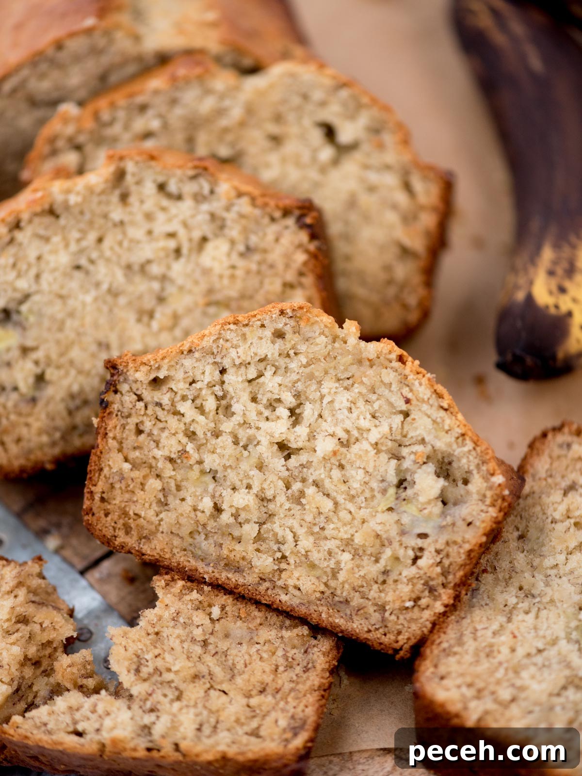 Classic Old-Fashioned Banana Bread Recipe for Perfect Loaf 5 Slices of freshly baked banana bread on a wooden surface, showcasing its moist and fluffy texture. A partially visible ripe banana is in the background, adding context to the flavor of the bread.