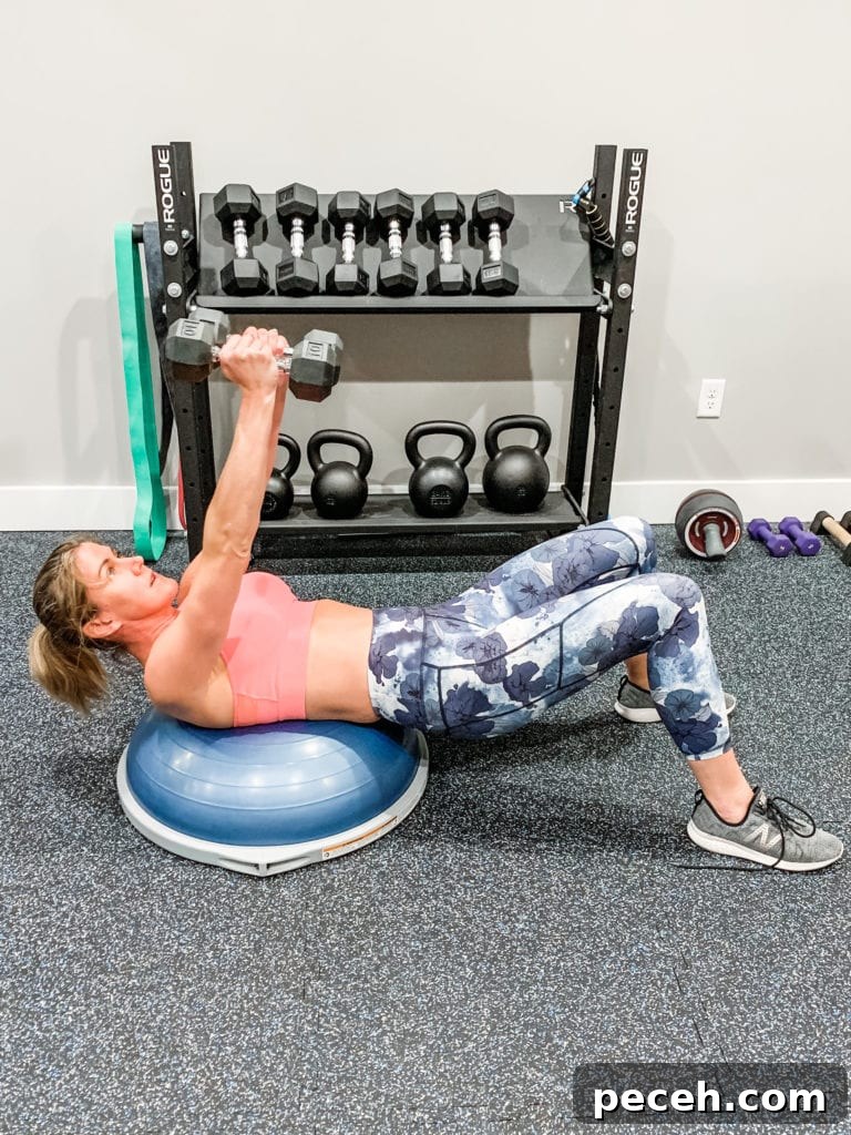 BOSU Ball Chest, Core & Quad Workout for Strength and Stability 5 a woman in floral leggings and a sports bra laying on a bosu ball and arms over her head with dumbbells