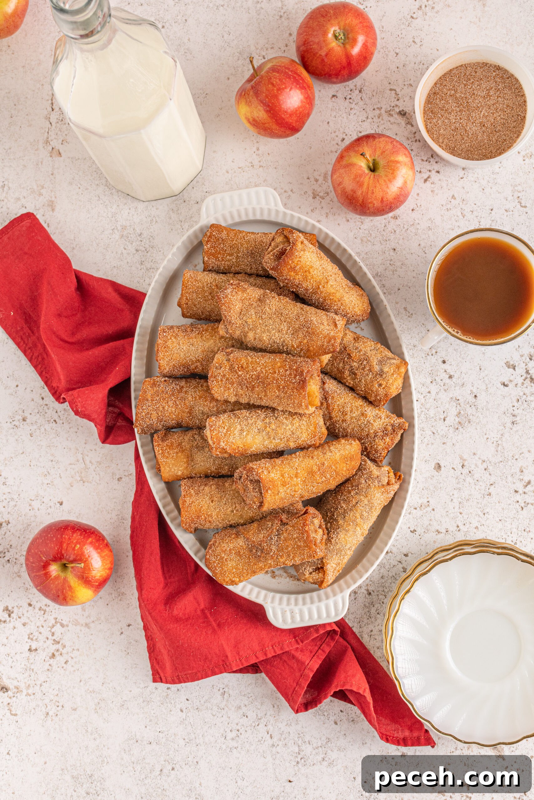 Crispy Apple Pie Egg Rolls with Cinnamon Sugar Dip 11 Overhead view of egg rolls stacked on a white platter with apples and a red linen in the background.