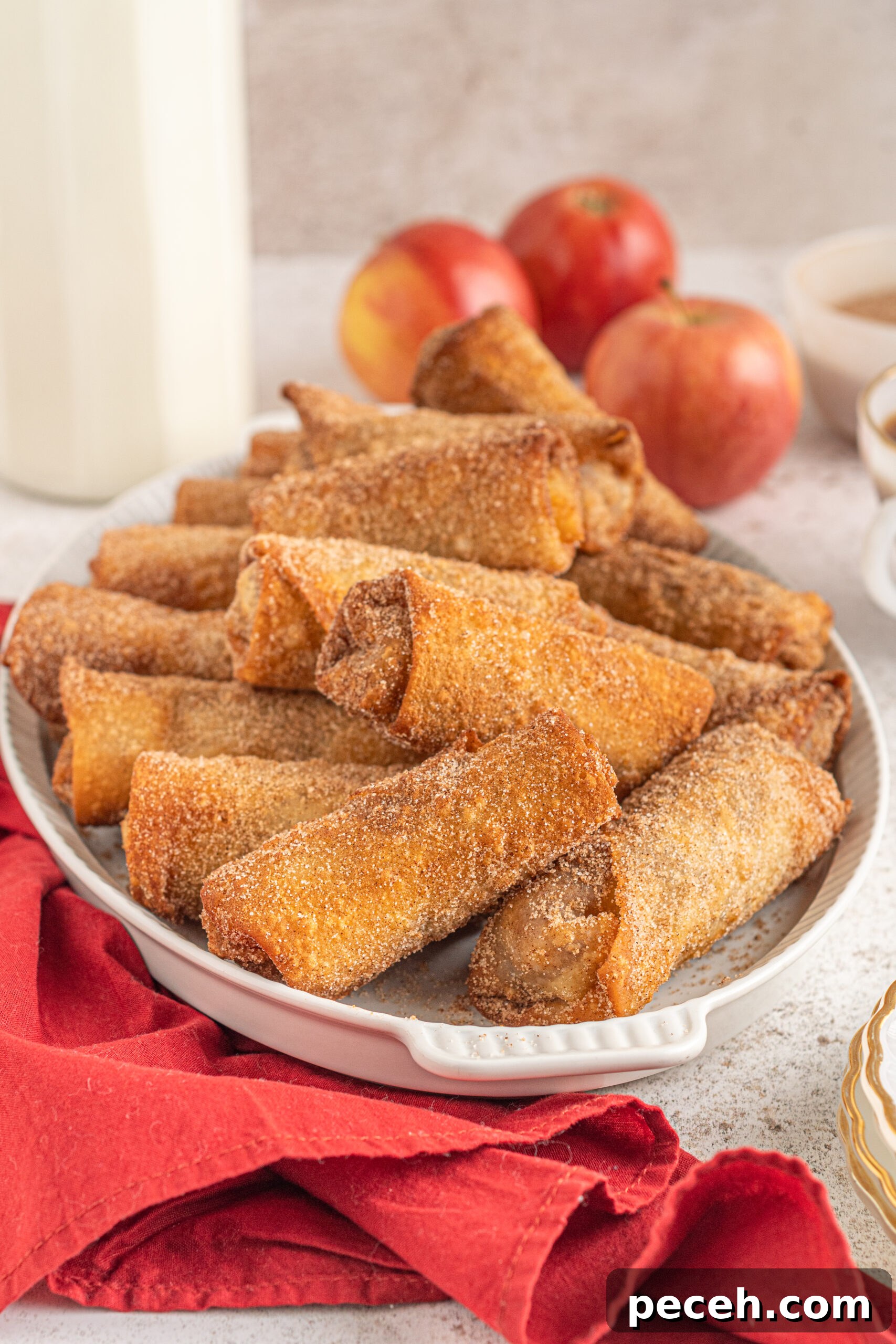Crispy Apple Pie Egg Rolls with Cinnamon Sugar Dip 3 Apple pie egg rolls stacked on a white platter with apples and a red linen in the background.