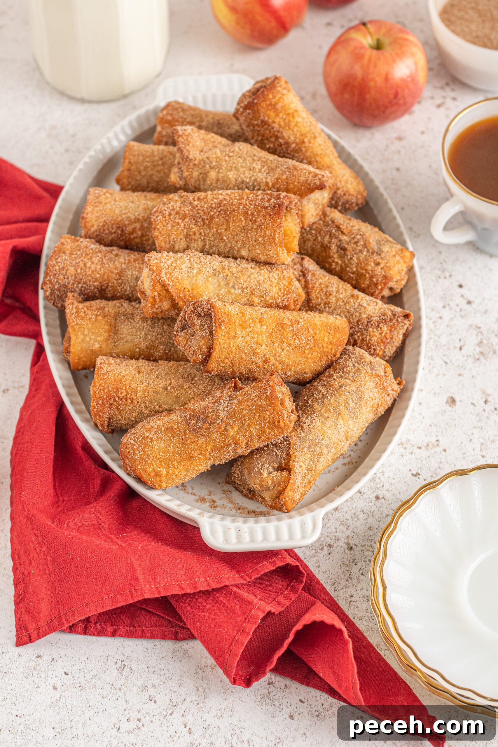 Crispy Apple Pie Egg Rolls with Cinnamon Sugar Dip 10 Egg rolls stacked on a white platter with apples and a red linen in the background.