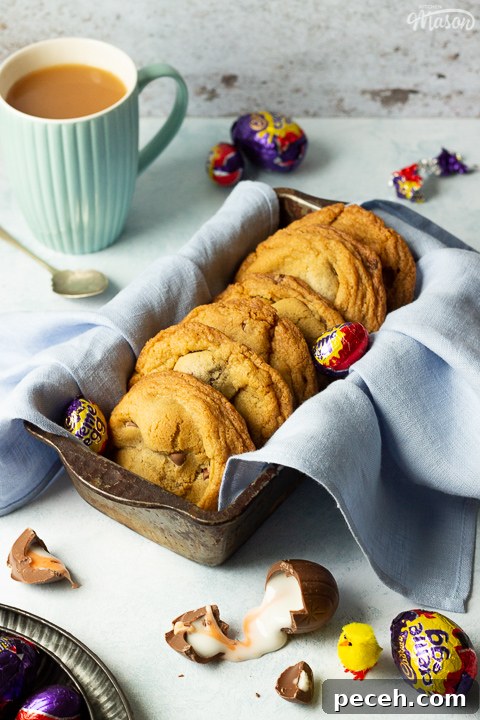 Crème Egg Cookie Recipe: Gooey Chocolate and Cadbury Filling 2 Creme Egg cookies in a bread tin lined with a pale blue napkin surrounded by whole and broken Creme Eggs. Set on a pale blue backdrop with a tray of whole Creme Eggs, a mug of tea and a yellow Easter chick in the background.
