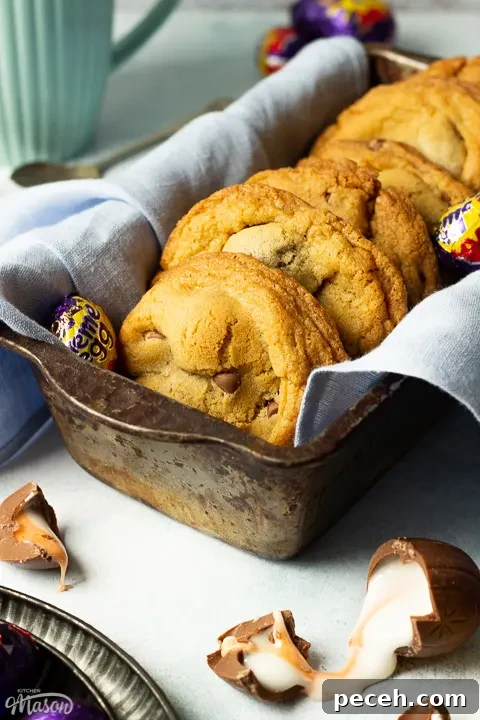 Crème Egg Cookie Recipe: Gooey Chocolate and Cadbury Filling 6 Creme Egg cookies in a bread tin lined with a pale blue napkin surrounded by whole and broken Creme Eggs. Set on a pale blue backdrop with a tray of whole Creme Eggs and a mug of tea in the background.
