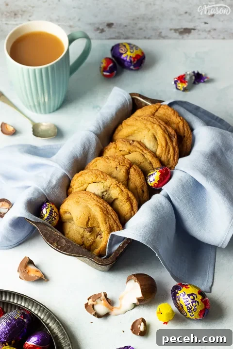 Crème Egg Cookie Recipe: Gooey Chocolate and Cadbury Filling 7 Creme Egg cookies in a bread tin lined with a pale blue napkin surrounded by whole and broken Creme Eggs. Set on a pale blue backdrop with a tray of whole Creme Eggs, a mug of tea and a yellow Easter chick in the background.