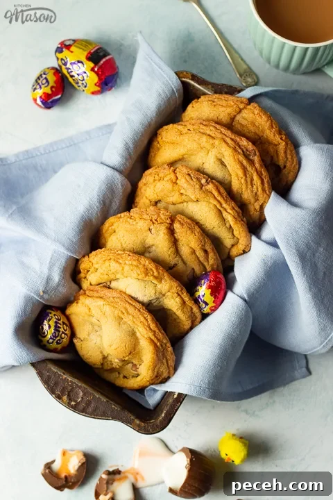 Crème Egg Cookie Recipe: Gooey Chocolate and Cadbury Filling 8 Birds eye view of Creme Egg cookies in a bread tin lined with a pale blue napkin surrounded by whole and broken Creme Eggs. Set on a pale blue backdrop with a mug of tea and a yellow Easter chick in the background.