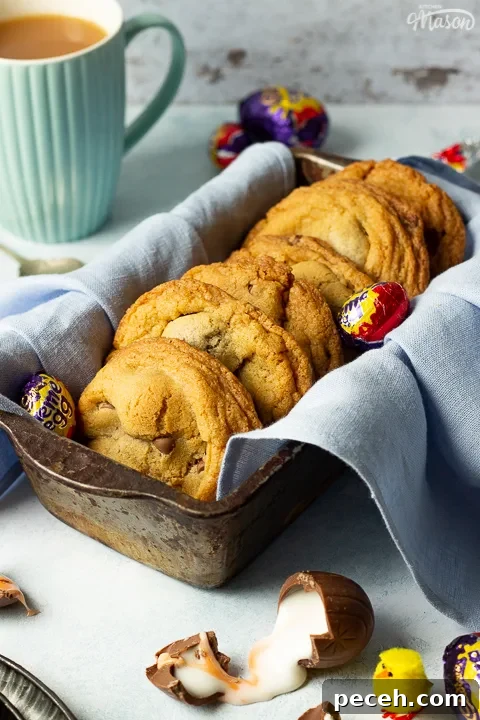 Crème Egg Cookie Recipe: Gooey Chocolate and Cadbury Filling 9 Creme Egg cookies in a bread tin lined with a pale blue napkin surrounded by whole and broken Creme Eggs. Set on a pale blue backdrop with a tray of whole Creme Eggs, a mug of tea and a yellow Easter chick in the background.