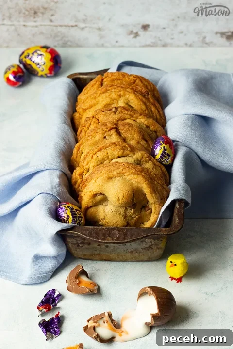 Crème Egg Cookie Recipe: Gooey Chocolate and Cadbury Filling 10 Front view of Creme Egg cookies in a bread tin lined with a pale blue napkin surrounded by whole and broken Creme Eggs. Set on a pale blue backdrop with a yellow Easter chick in the background.