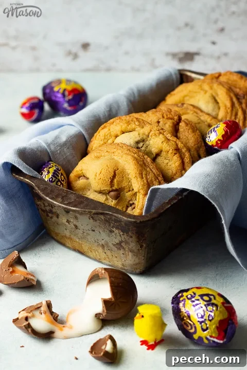 Crème Egg Cookie Recipe: Gooey Chocolate and Cadbury Filling 11 Creme Egg cookies in a bread tin lined with a pale blue napkin surrounded by whole and broken Creme Eggs. Set on a pale blue backdrop with a yellow Easter chick in the background.