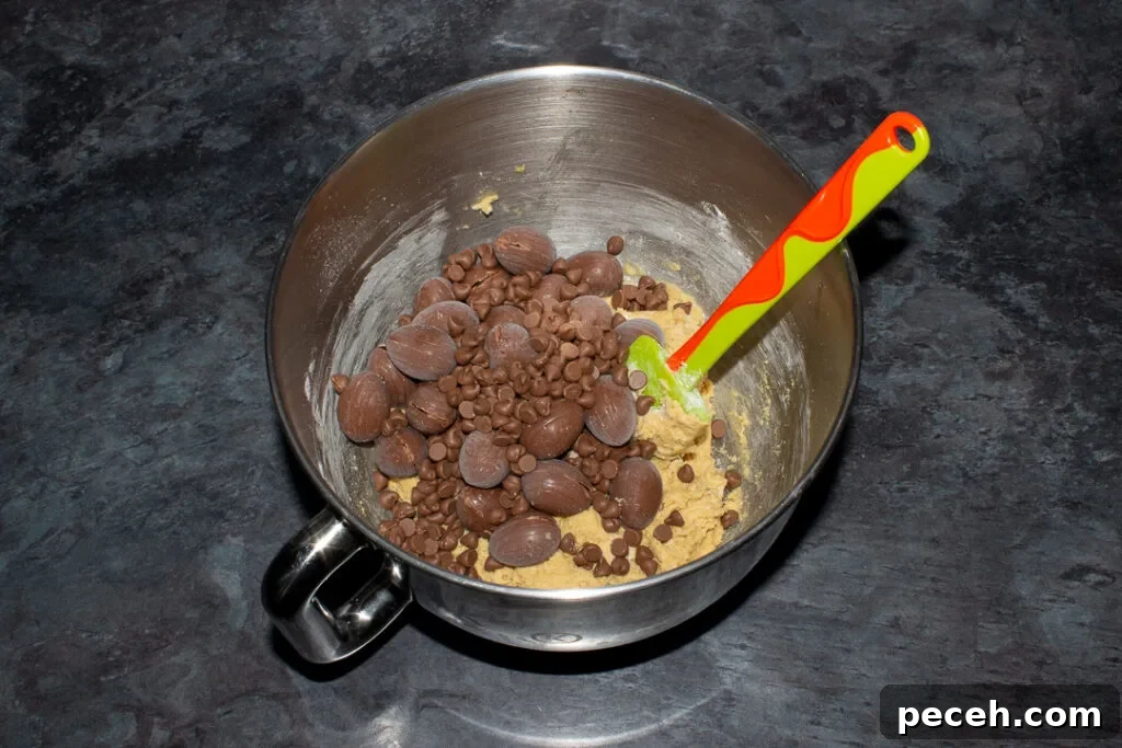 Crème Egg Cookie Recipe: Gooey Chocolate and Cadbury Filling 15 Frozen Creme Eggs and chocolate chips being added to cookie dough in the bowl of an electric stand mixer with a green spatula