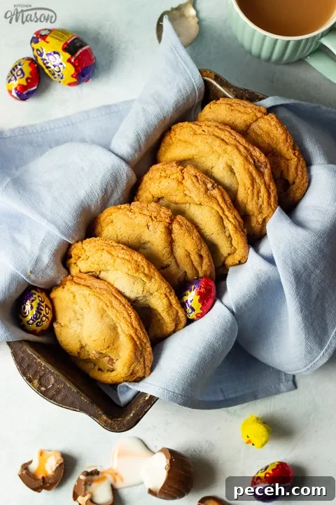 Crème Egg Cookie Recipe: Gooey Chocolate and Cadbury Filling 3 Birds eye view of Creme Egg cookies in a bread tin lined with a pale blue napkin surrounded by whole and broken Creme Eggs. Set on a pale blue backdrop with a mug of tea and a yellow Easter chick in the background.