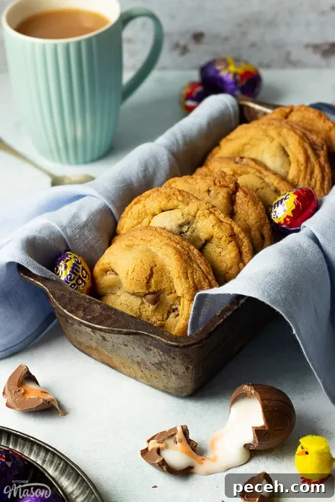 Crème Egg Cookie Recipe: Gooey Chocolate and Cadbury Filling 20 Creme Egg cookies in a bread tin lined with a pale blue napkin surrounded by whole and broken Creme Eggs. Set on a pale blue backdrop with a tray of whole Creme Eggs, a mug of tea and a yellow Easter chick in the background.