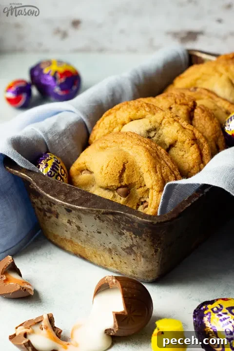 Crème Egg Cookie Recipe: Gooey Chocolate and Cadbury Filling 22 Creme Egg cookies in a bread tin lined with a pale blue napkin surrounded by whole and broken Creme Eggs. Set on a pale blue backdrop with a yellow Easter chick in the background.