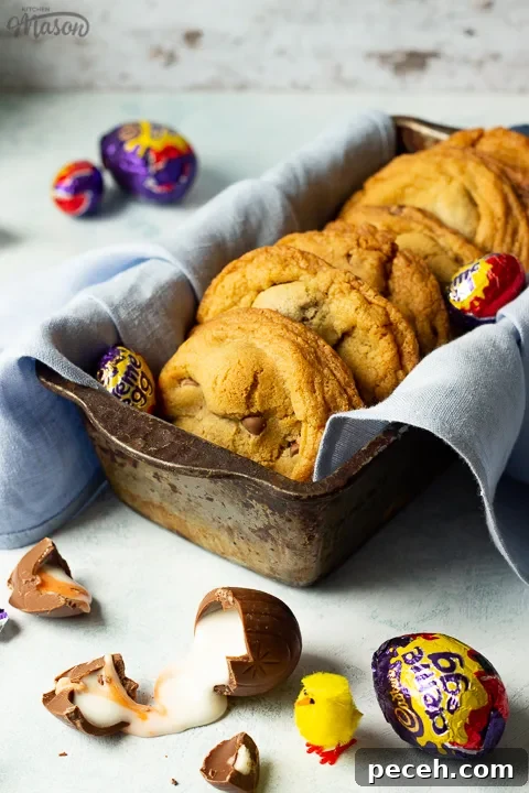 Crème Egg Cookie Recipe: Gooey Chocolate and Cadbury Filling 4 Creme Egg cookies in a bread tin lined with a pale blue napkin surrounded by whole and broken Creme Eggs. Set on a pale blue backdrop with a yellow Easter chick in the background.