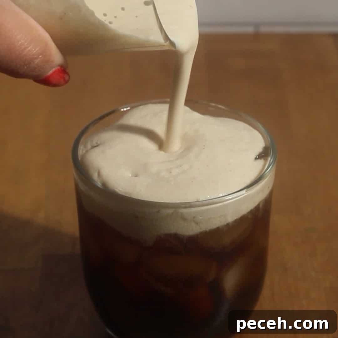 Pumpkin cold foam being poured over iced coffee in a clear glass.