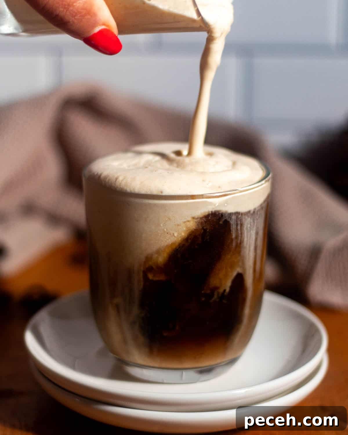 Pumpkin cold foam being poured over a glass of iced coffee with ice cubes visible.