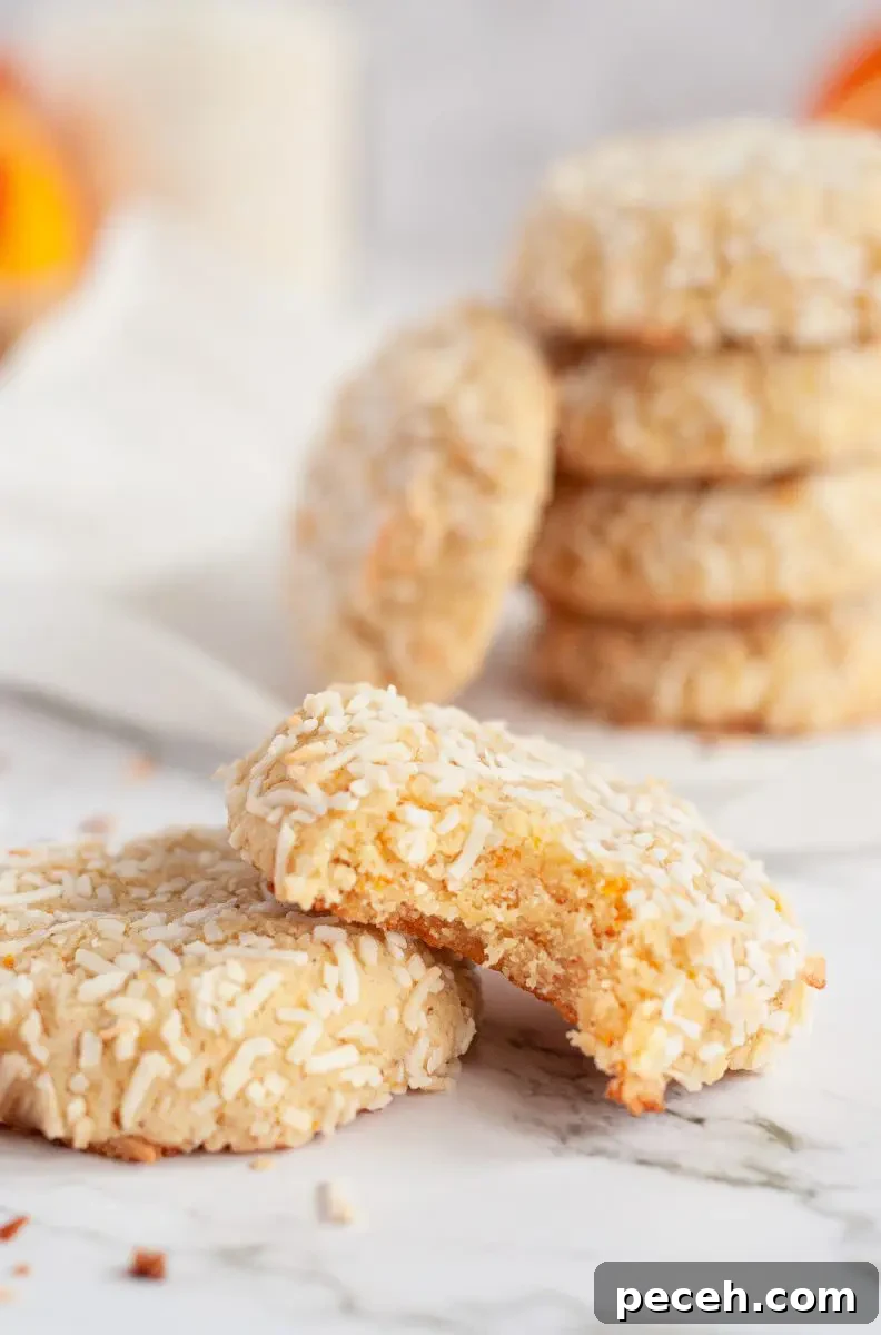 Coconut orange cookies covered in white shredded coconut. There are two cookies sitting in front of a cookie stack and one of the cookies is leaning up against the other cookie and has a large bite taken out of it.
