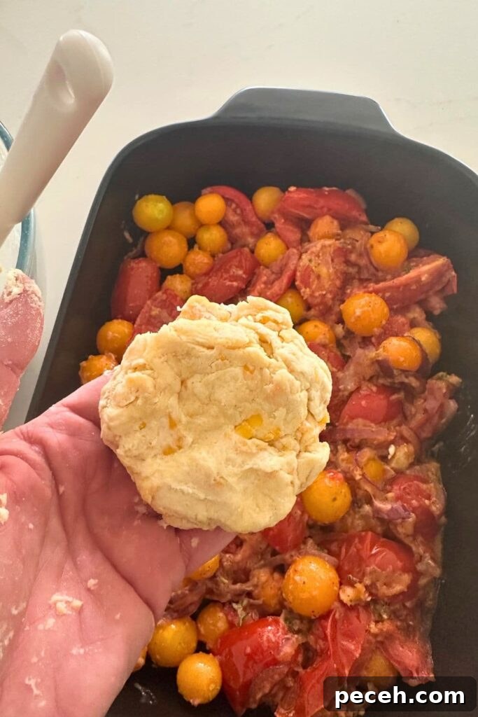 A photo of the base of a sourdough tomato cobbler in a black baking dish. There a hand holding the sourdough cheddar biscuit topping as it's being added to the base.