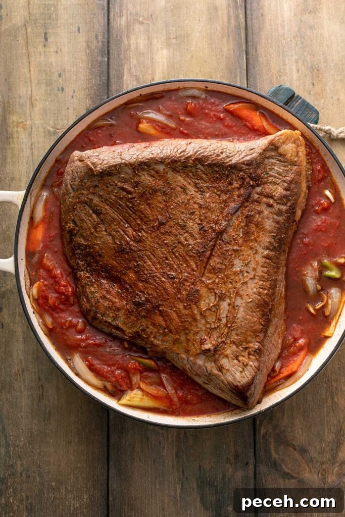Seared beef brisket in a pan on a bed of vegetables and sauce about to be put in the oven for braising.