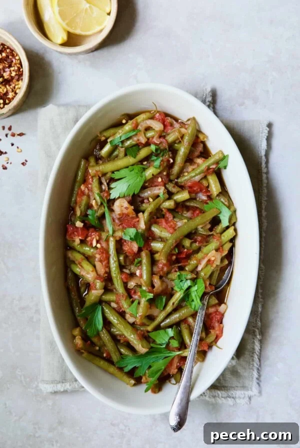 Mediterranean Braised Green Beans with Tomatoes in oval serving dish with serving spoon garnished with fresh parsley.