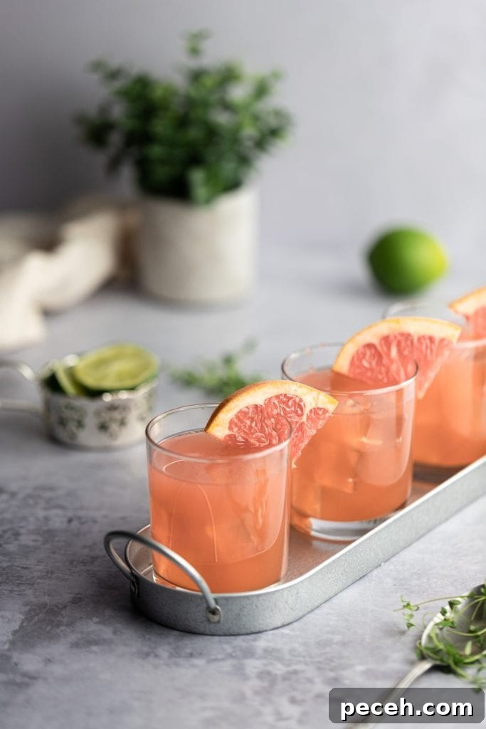 Sparkling Grapefruit Crush: Refreshing Citrus Cocktail Recipe 2 Three grapefruit crush cocktails garnished with grapefruit slices lined up in a row on an angle in a shiny metal tray, limes in the background.
