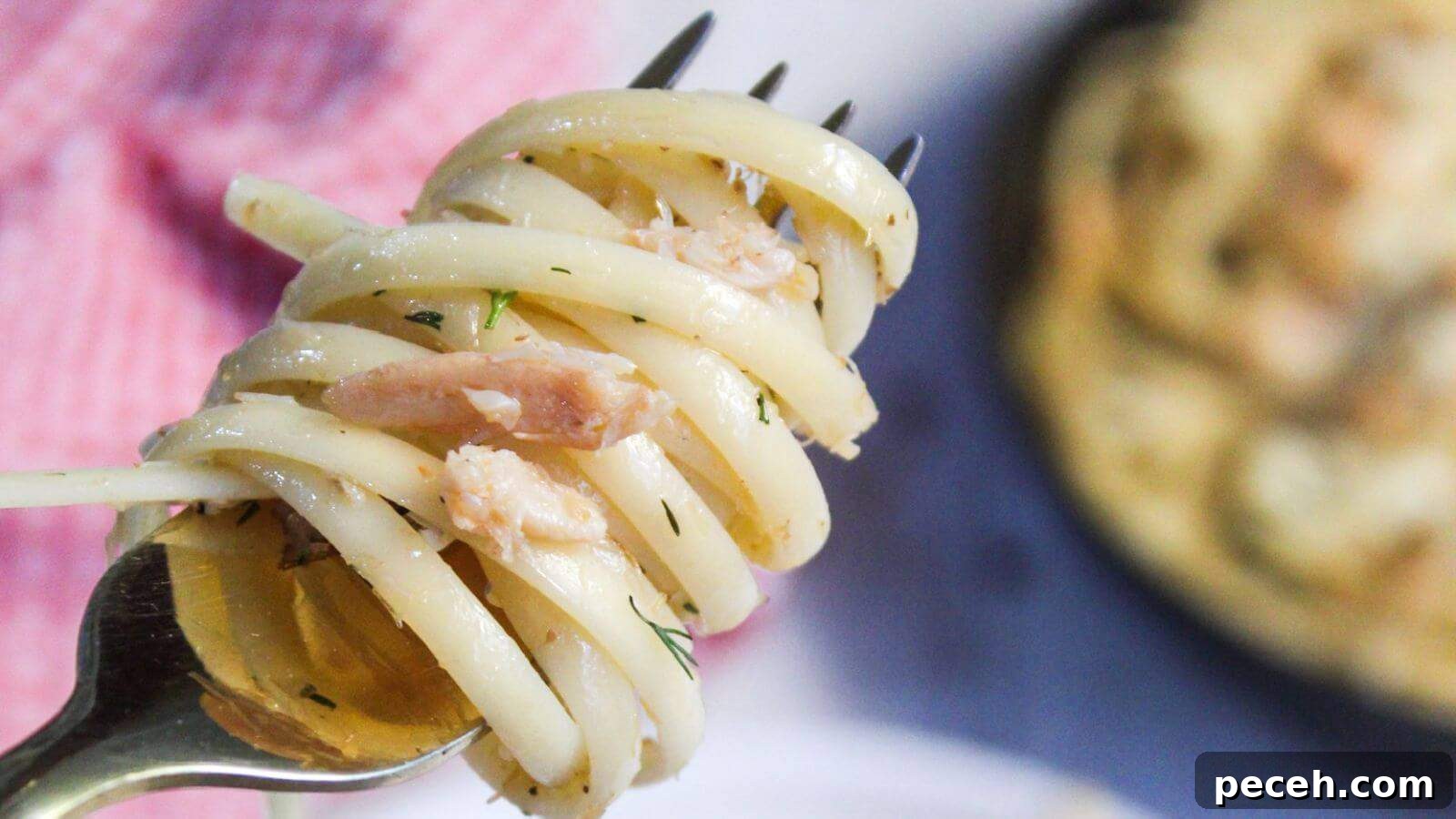 A fork twirling crab pasta with bits of herbs, set against a plate with more pasta and a blurred background.