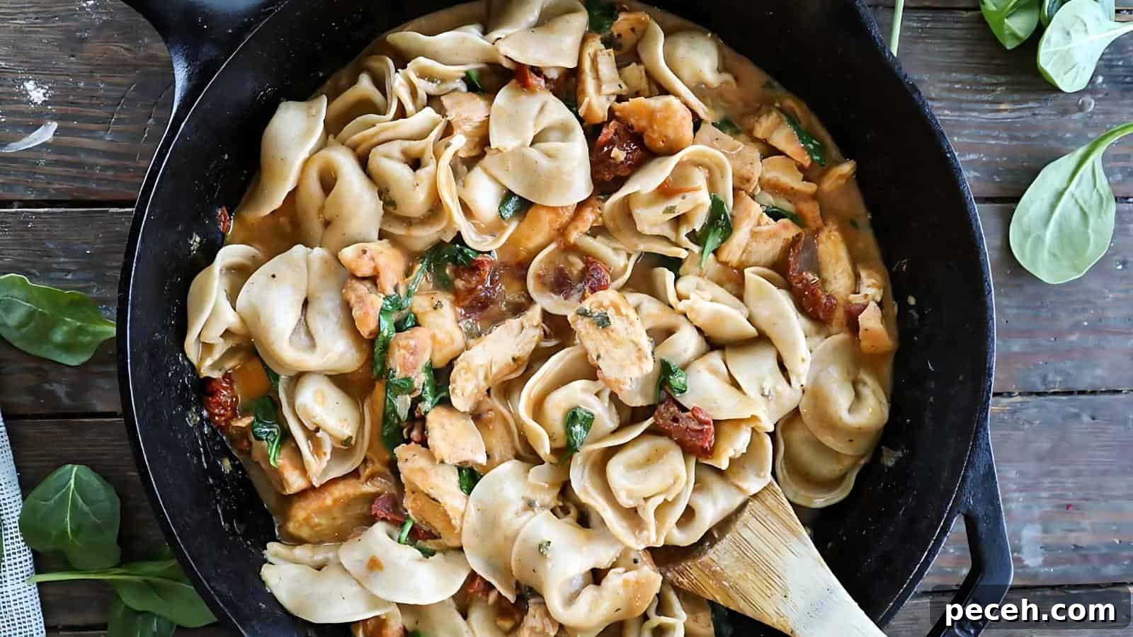 A skillet filled with pasta, pieces of chicken, sun-dried tomatoes, and spinach in a creamy sauce, with a wooden spatula on the side.