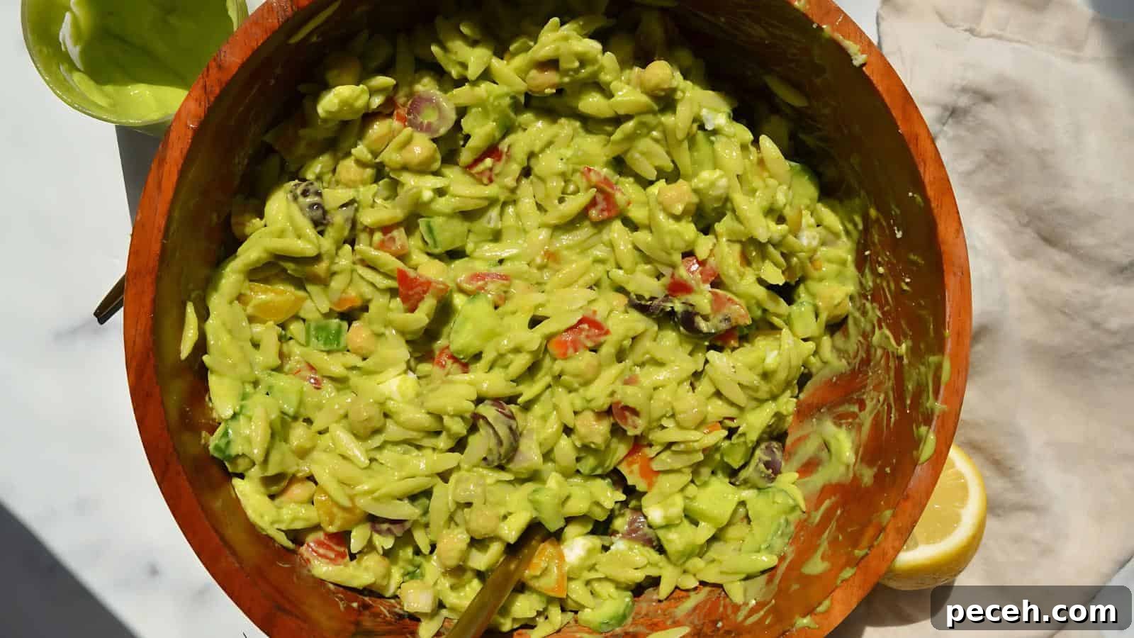 A wooden bowl filled with a creamy green pasta salad, mixed with chopped vegetables, beside a cut lemon and a green sauce.