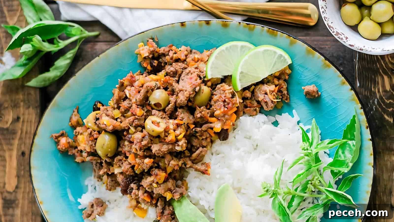A dish with seasoned ground meat and green olives on a blue plate, served with white rice, lime wedges, and fresh herbs.