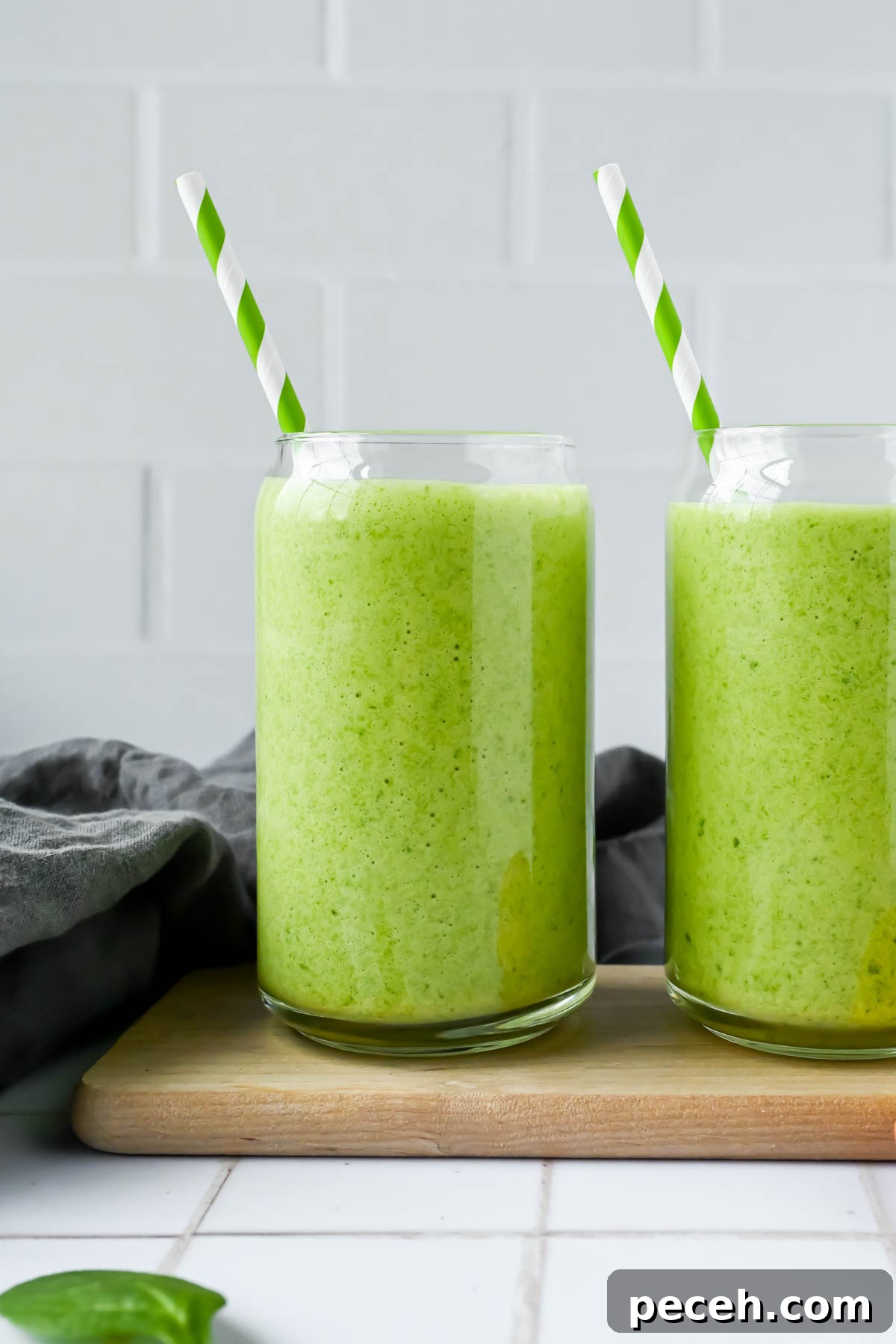 Mango Spinach Smoothie Recipe for Energy and Glow 2 two mango spinach smoothies with a green and white striped straw on white tile background.