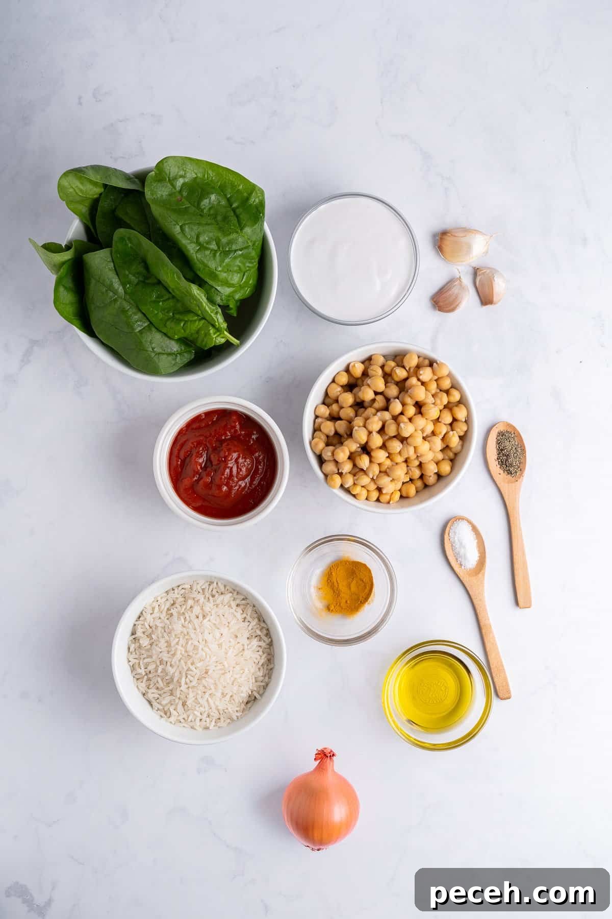 Ingredients laid out on a white surface like: fresh spinach, garlic cloves, tomato paste, uncooked rice, onion, and small bowls of olive oil, turmeric, pepper, and salt.