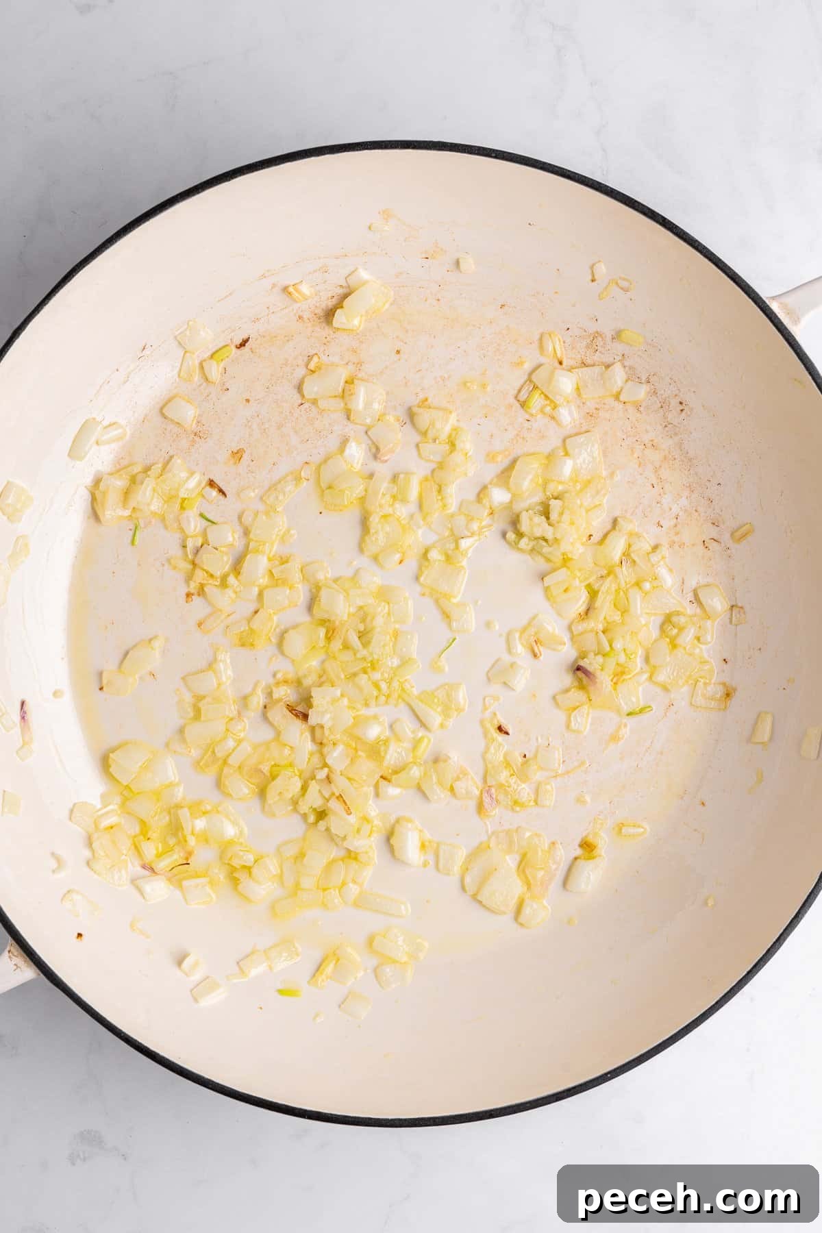 Chopped onions sautéing in a white pan on a marble surface. The onions are slightly golden, indicating they are being cooked. The pan shows hints of browning, suggesting the onions are being caramelized.