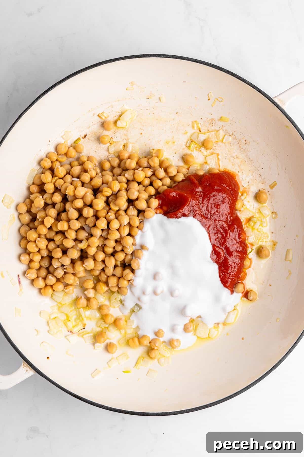 A white pot containing cooked chickpeas, chopped onions, tomato sauce, and coconut milk, ready to be mixed.